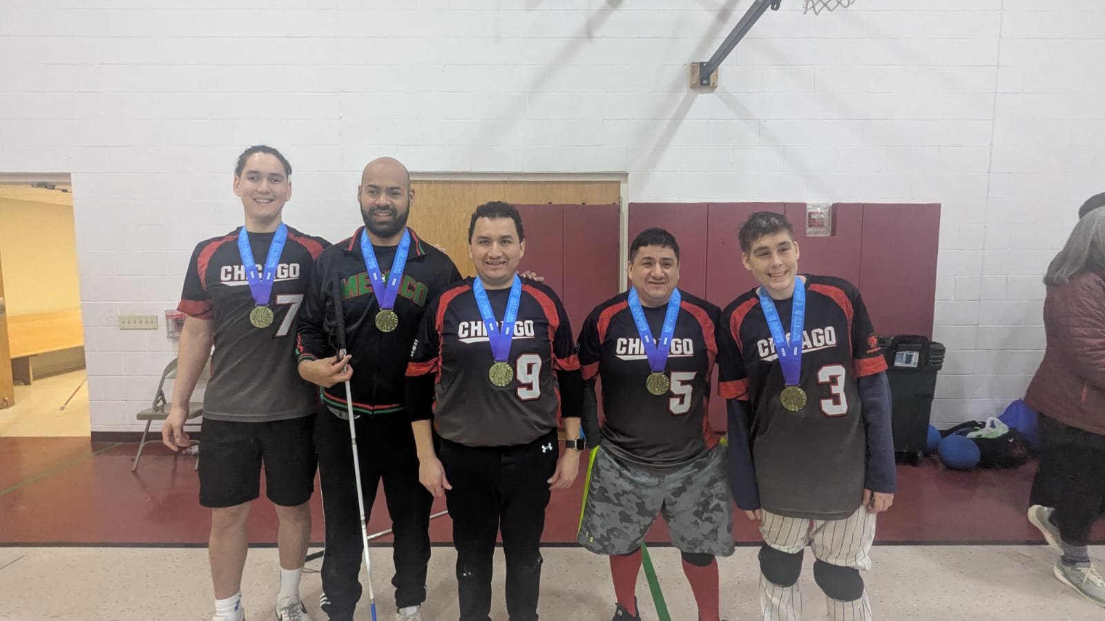 Chicago Fury men's team from left to right: Ardan- 7, Diego -8, Juan -9, Oscar -5, and Mike -3. All men are wearing the red and black jersey which reads Chicago in white with their respective jersey numbers and all smiling. They have gold medals on which have blue ribbons. There is a white wall with a red mat in the background and is taken inside of a gymnasium. There are goalballs on the right side in the floor behind them